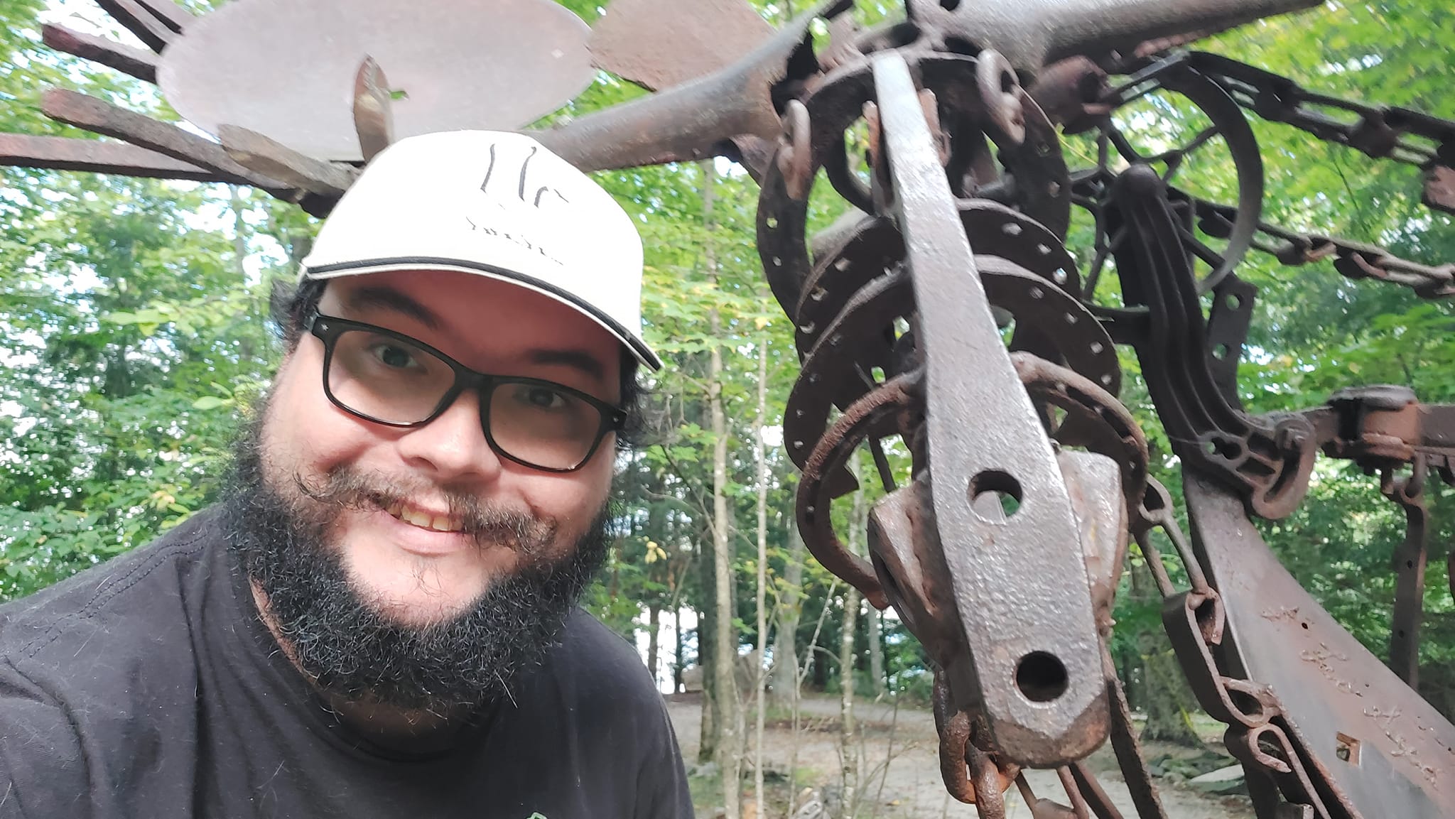 A man with short hair and bushy facial hair stands in front of a sculpture of a moose made of scrap metal pieces, several horseshoes and rail road spikes are able to be made out